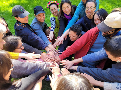 16 participants from the 2024 Asian Diaspora jam are in a circle outside, smiling with their hands in the center in an ‘all in’ moment
