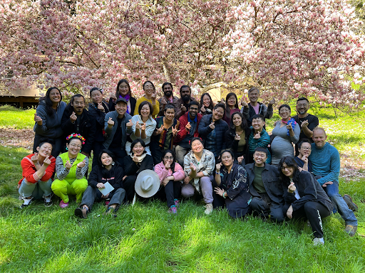 29 participants of the 2024 Asian Diaspora Jam gather outside on the grass under a Cherry Blossom tree. There are 3 rows of people, some standing, some kneeling, some finger hearting, all smiling, in close warm proximity to each other,